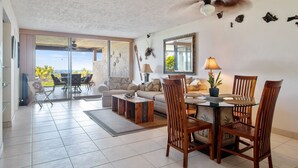 A spacious living area with a dining table, chairs, and a sofa. There is a large window offering a view of palm trees.