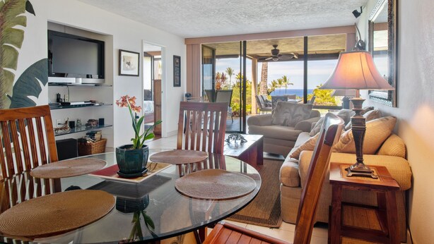 A modern living room with a glass dining table, comfortable seating, a television, and a view of the outdoors.