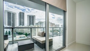 A balcony with a view of high-rise buildings, a seating area with cushions, and a pool in the background. - New luxurious 3bdr condo at BW with Resort amenities & Beach Chairs  (Hallandale Beach)