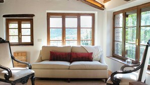 A room with a wooden ceiling, a sofa with a red and white checkered pillow, a chair, and a window with a view of greenery.