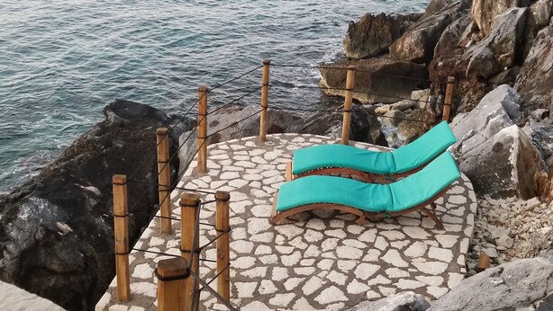 Un espace extérieur aménagé avec un sol en pierre, comprenant un banc en bois et un coussin turquoise, offrant une vue sur une côte rocheuse et la mer.