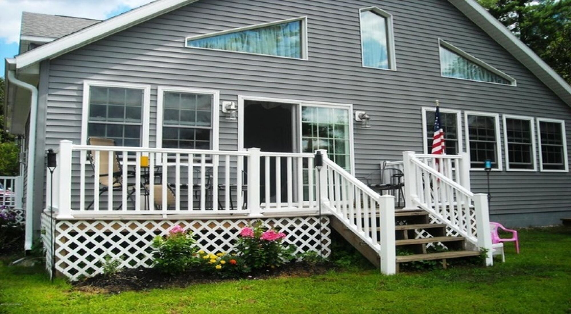 A two-story house with a front porch, a small garden with flowers, and an American flag.