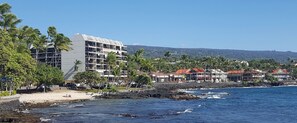 A coastal hotel with multiple floors, a beach in the foreground, and a view of the ocean and distant hills.