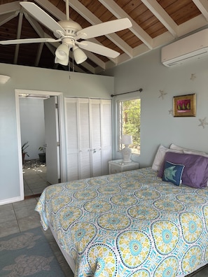 A bedroom with a patterned bedspread, a ceiling fan, a window with a view, and a door leading to an outdoor area.