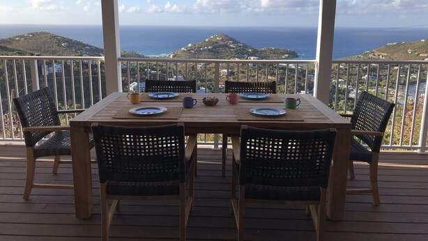 Une terrasse en bois avec une table dressée pour deux personnes, offrant une vue sur un paysage côtier avec des collines et la mer.