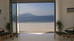 Et moderne værelse med en stor skydedør, der fører ud til en balkon med udsigt over havet og fjerne bjerge. Der er to træstole og en potteplante på balkonen.