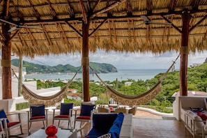 Una silla hamaca bajo un techo de paja, con vista al mar, y un conjunto de mesa y sillas.