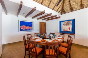 Un comedor con una mesa redonda dispuesta para seis personas, rodeada de sillas de madera con cojines naranja. Hay dos obras de arte enmarcadas en las paredes, una puerta de madera y un techo con vigas a la vista.