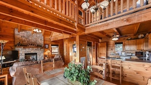 Interior acolhedor de uma cabana, com lareira de pedra, mesa de jantar de madeira e cozinha.