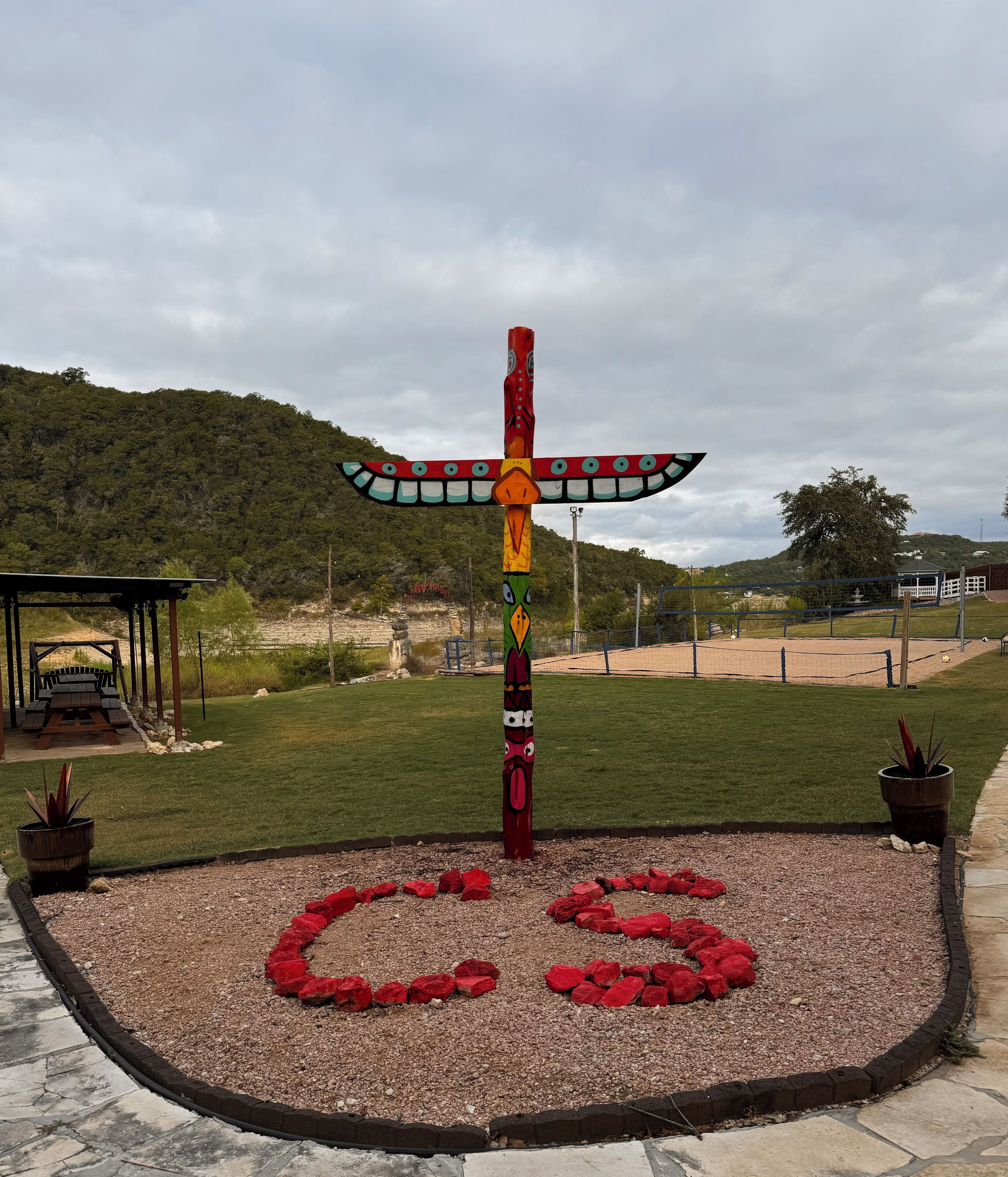 A totem pole with a cross design, surrounded by a gravel area with red flowers, set against a backdrop of a grassy field and a forested hill.