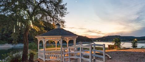 A gazebo with a wooden deck, surrounded by a gravel path and pink flowers, overlooking a body of water and hills.