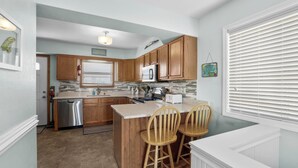 A kitchen with wooden cabinets, a stainless steel dishwasher, a refrigerator, and a countertop with two wooden chairs.
