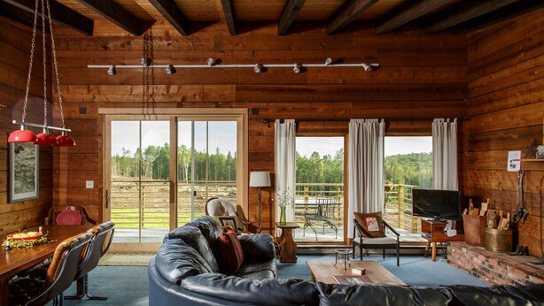A cozy cabin interior with a leather sofa, dining table, and chairs, wooden walls, and large windows offering a view of the outdoors. - Reillys Rest-private home in Green Mtns, kid-pet friendly-Ski,Bike,Walk,Relax! (Weston)