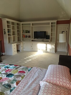 Ein Schlafzimmer mit einem großen Bett, einem an einer Wand festmontierten Fernseher und einem Fenster, durch das natürliches Licht einfällt.
