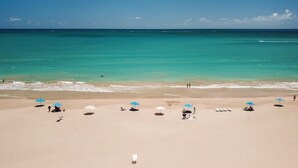 Ein Strand mit klarem türkisblauem Wasser, mehreren Sonnenschirmen und Menschen, die die Sonne genießen.