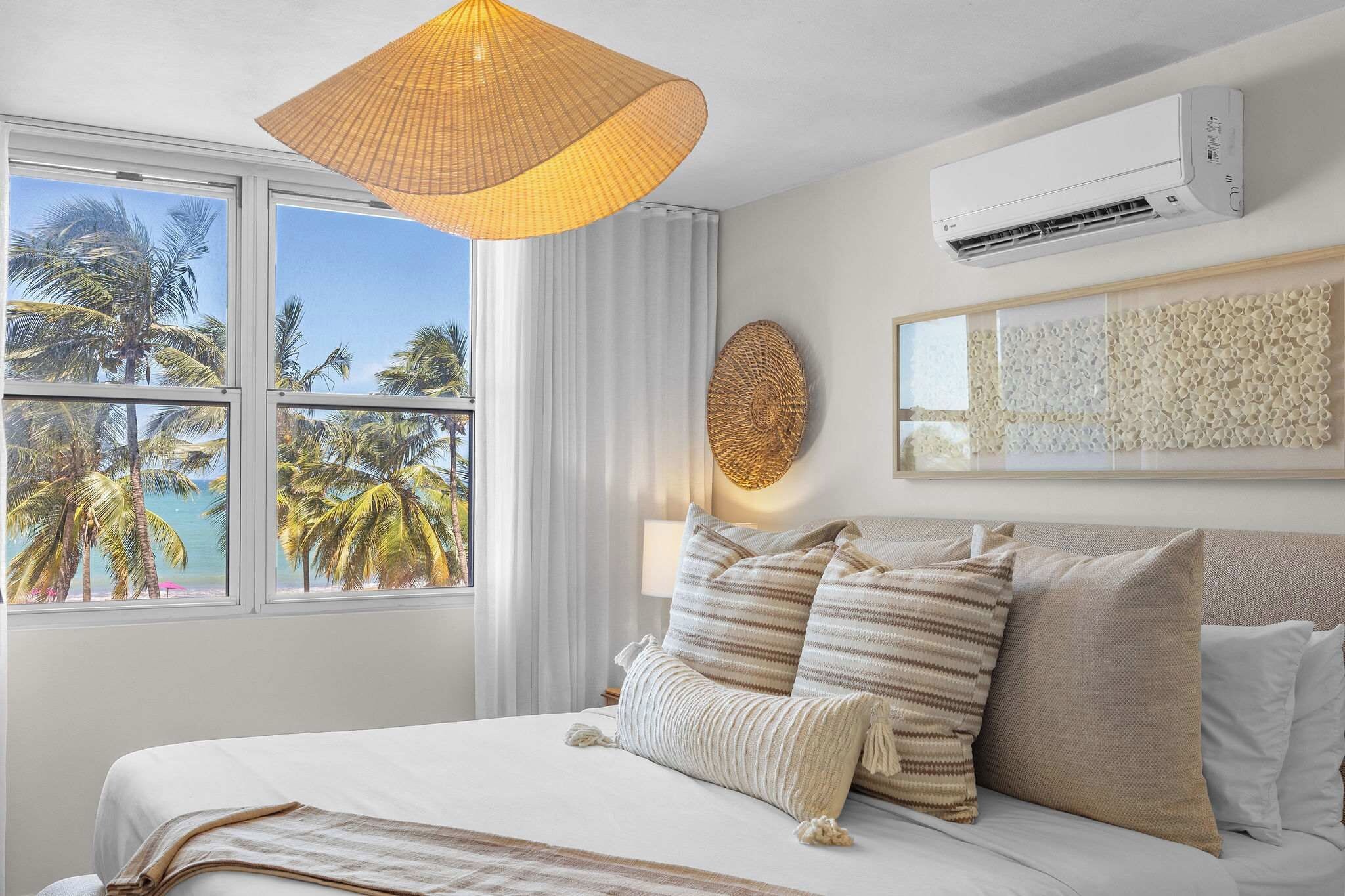 Quarto de hotel com cama, uma janela grande com vista para palmeiras e o mar, ar-condicionado fixado na parede e uma obra de arte com textura na parede.