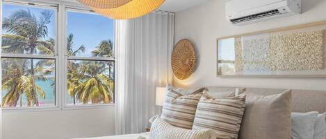 A hotel room with a bed, a large window with a view of palm trees and the sea, a wall-mounted air conditioner, and a textured wall art piece.