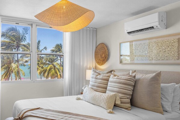 A hotel room with a bed, a large window with a view of palm trees and the sea, a wall-mounted air conditioner, and a textured wall art piece.