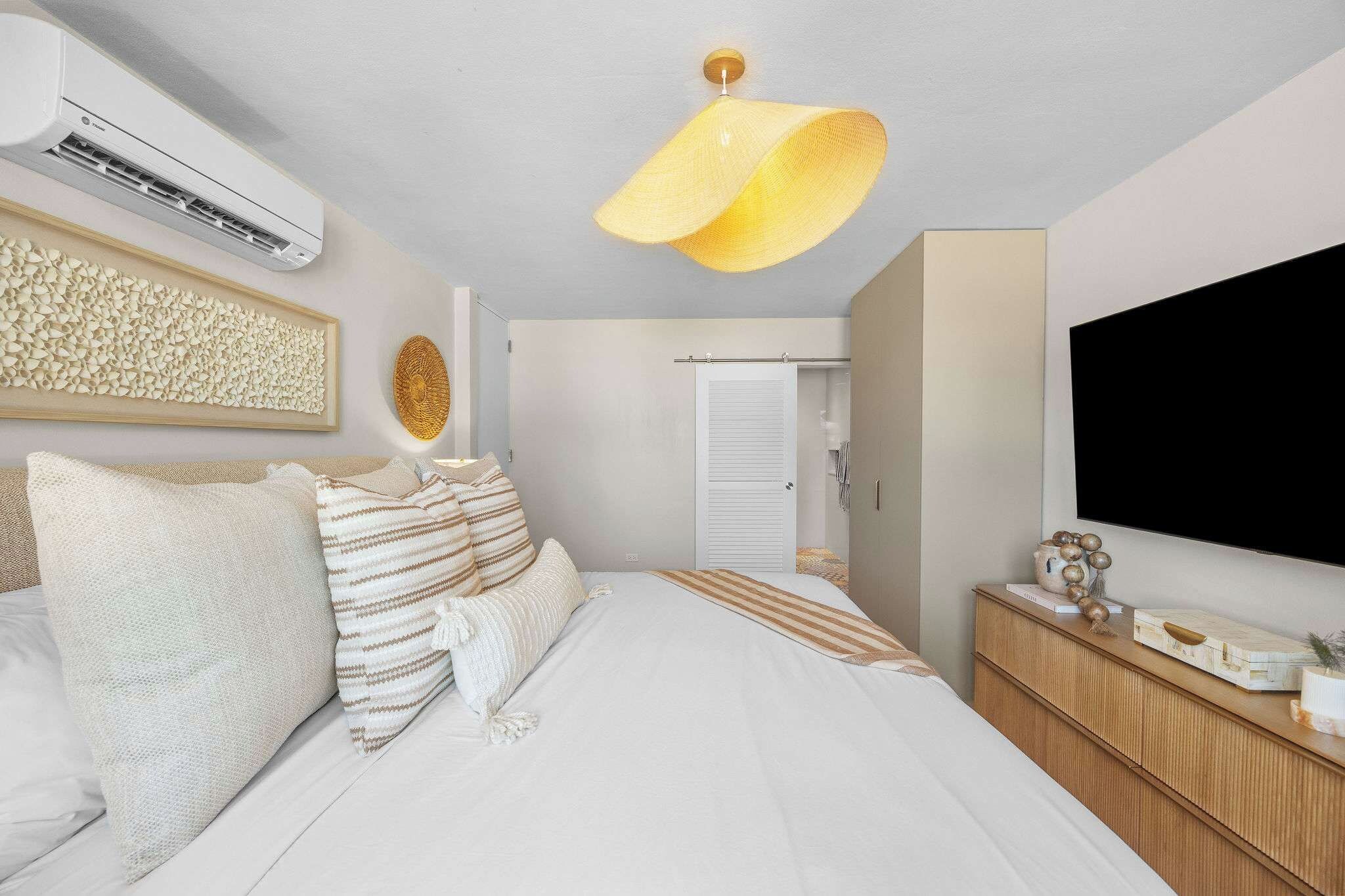 A bedroom with a bed, pillows, a bedside table, a TV, and an air conditioning unit.