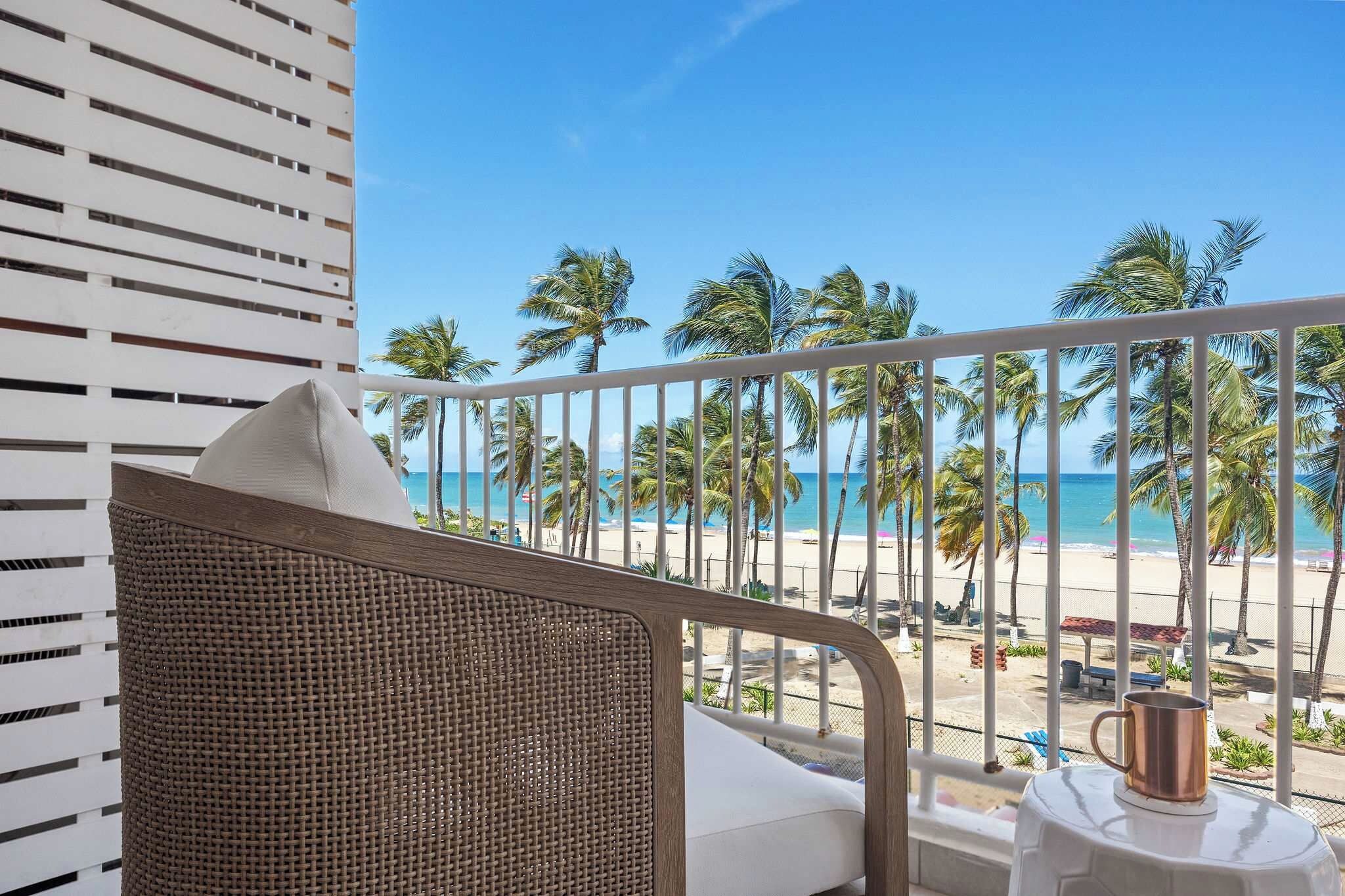 A balcony with a wicker chair and a metal railing overlooking a beach with palm trees and the ocean.