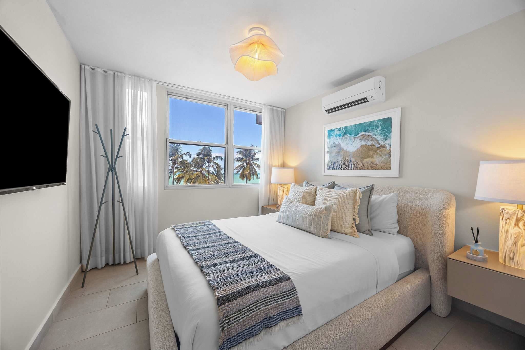Una habitación de hotel con una cama, un ventanal con vistas a palmeras, un aire acondicionado instalado y un cuadro en la pared.