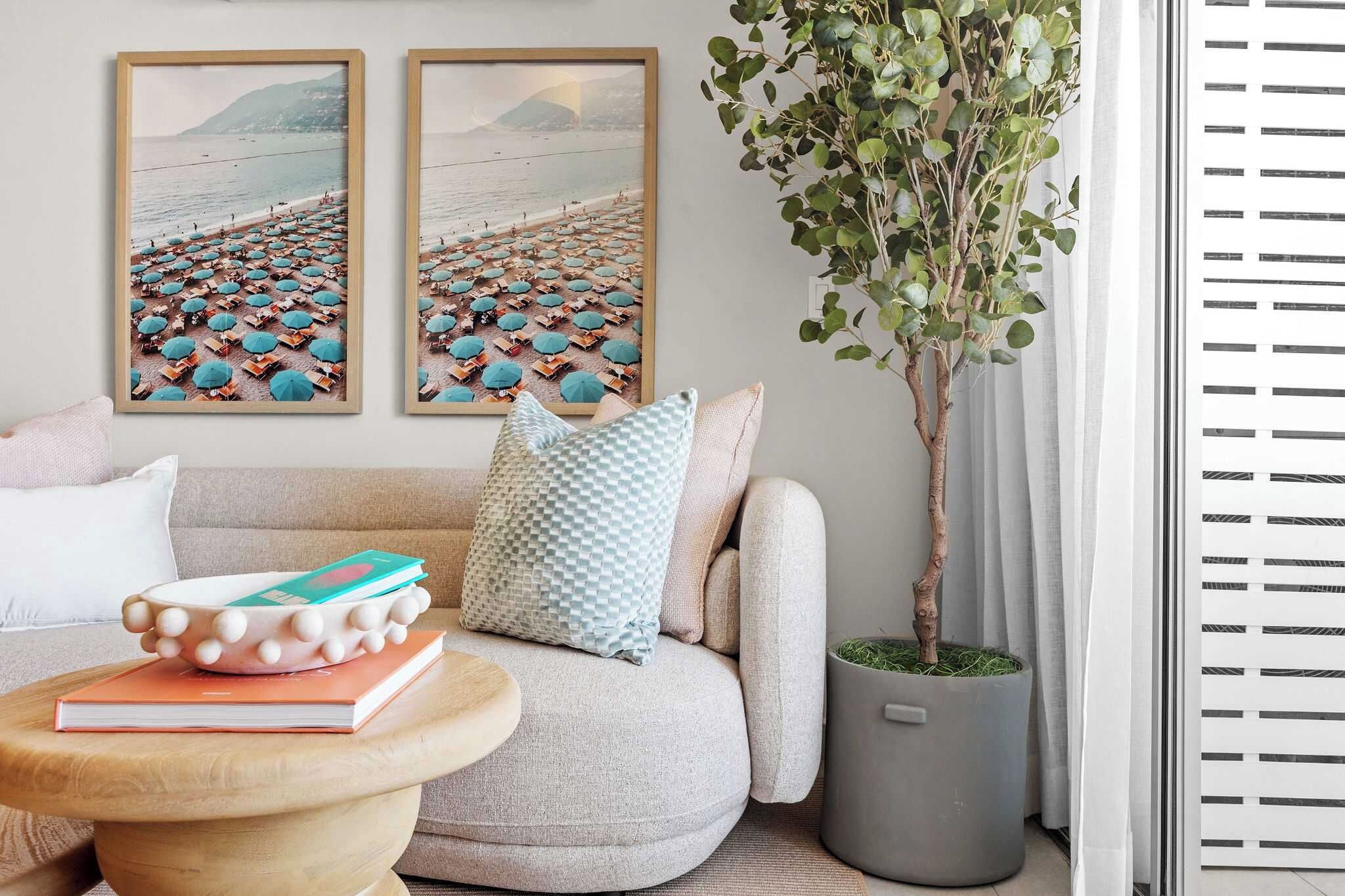 A living room with a sofa, a coffee table with books, a potted plant, and two beach-themed wall art pieces.