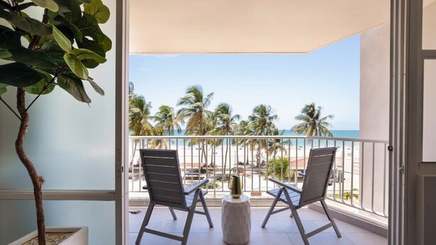 Ein Balkon mit zwei Stühlen und einem kleinen Tisch, mit Blick auf einen Strand mit Palmen und dem Meer.