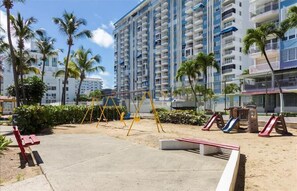 Un parque infantil con columpios y toboganes, rodeado de palmeras y un edificio alto al fondo.