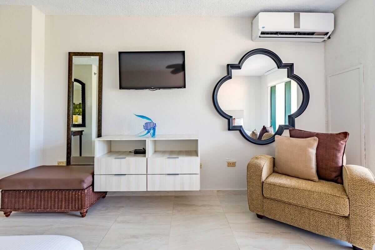 Un soggiorno moderno con TV a schermo piatto, un grande specchio decorativo, un poggiapiedi in vimini e un divano color beige.