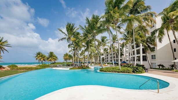 Piscina de resort con palmeras, playa y cielo despejado.