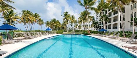 A swimming pool with lounge chairs and umbrellas, surrounded by palm trees and a multi-story building.