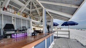 Un bar situé en bord de mer, avec un comptoir en bois, des étagères remplies de bouteilles et des parasols.
