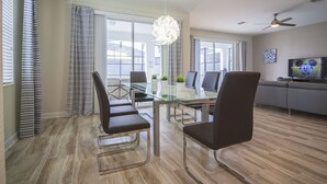 Un espace repas moderne avec une table en verre et des chaises en métal, de grandes fenêtres ornées de rideaux à rayures, un ventilateur de plafond et un téléviseur diffusant un dessin animé.