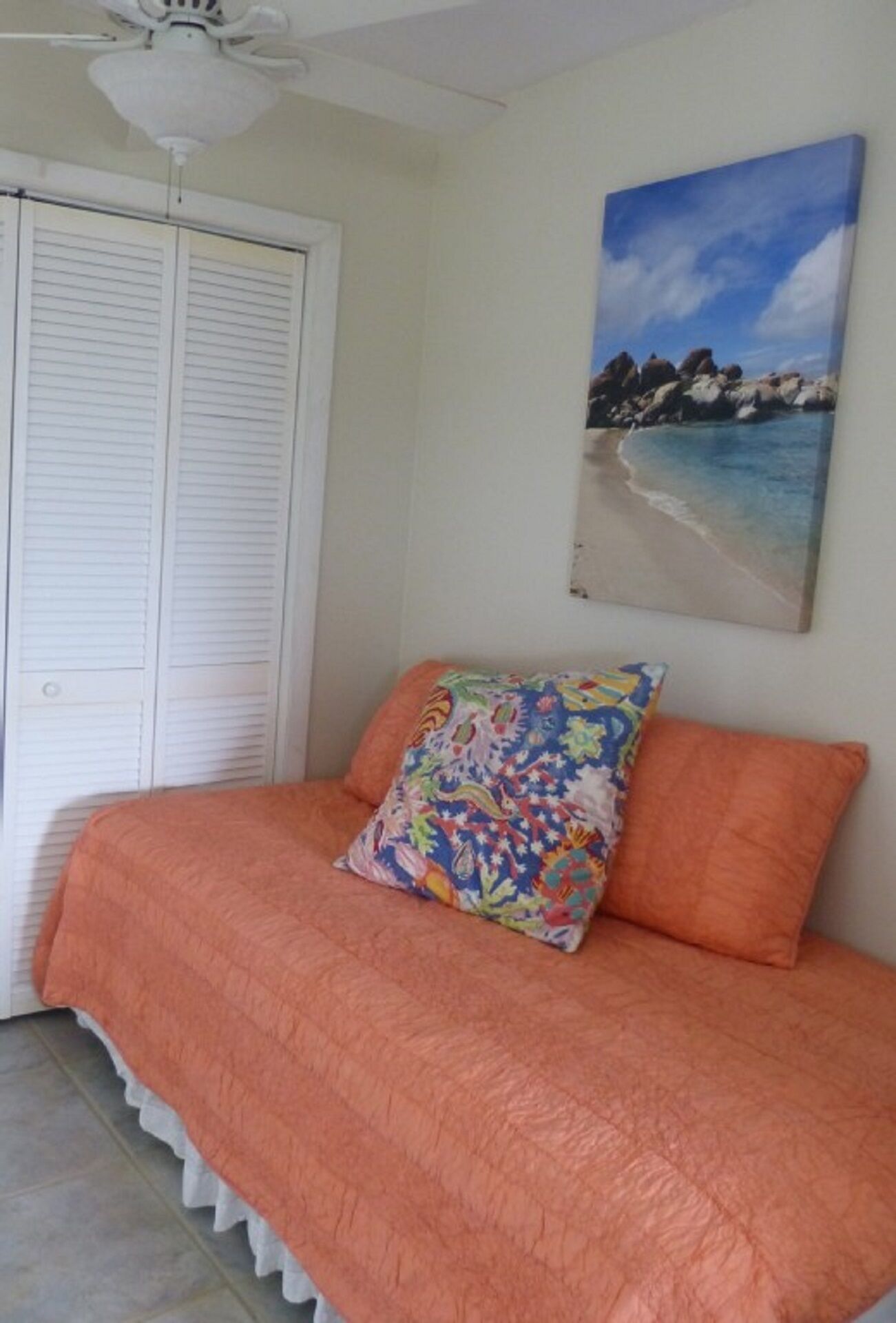 Un dormitorio con cama, colcha naranja, almohada colorida, ventilador de techo y un cuadro con una escena de playa.