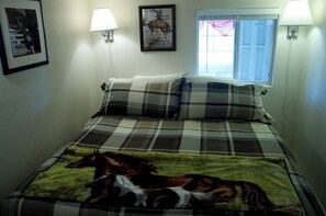 Une chambre à coucher avec un lit recouvert d’une couvre-lit à motifs équestres, deux lampes fixées au mur, une affiche encadrée et une fenêtre avec des rideaux.