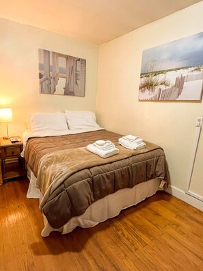 A neatly made bed with a brown comforter and white pillows, a nightstand with a lamp, and two wall-mounted paintings depicting architectural and beach scenes. - 1st Base Apt 1 - All Star Baseball Rentals (Oneonta)