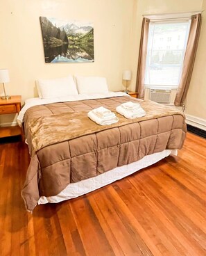 A neatly made bed with a brown quilt, two folded white towels, a nightstand with a lamp, a wall-mounted painting of a mountain landscape, and a window with curtains. - 1st Base Apt 1 - All Star Baseball Rentals (Oneonta)
