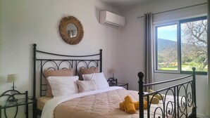 Un lit bien fait, recouvert d’une couette beige, une table de chevet avec une lampe, et une vue sur l’extérieur par une fenêtre.