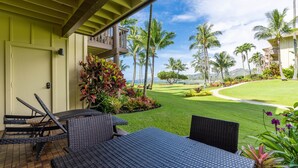 A covered outdoor seating area with wicker furniture, a view of a well-maintained lawn, and tall palm trees.