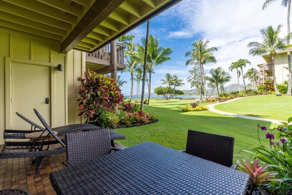 A covered outdoor seating area with wicker furniture, a view of a well-maintained lawn, and tall palm trees.