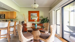 A dining area with wicker chairs and a glass table, a kitchen with wooden cabinets, and a large window with blinds.