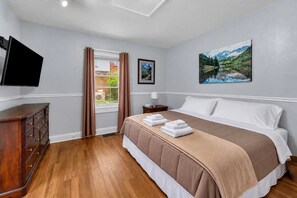 Hotellihuone, jossa on sänky, televisio, lipasto, ikkuna verhoilla ja taulu seinällä.