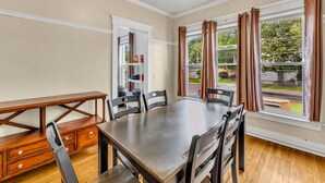 A dining area with a wooden table and chairs, a built-in bookshelf, and large windows with curtains. - 2nd Base Apt 1 - All Star Baseball Rentals (Oneonta)