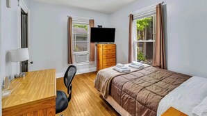 A hotel room with a bed, a desk with a lamp, a TV on a dresser, and a window with curtains. - 2nd Base Apt 1 - All Star Baseball Rentals (Oneonta)