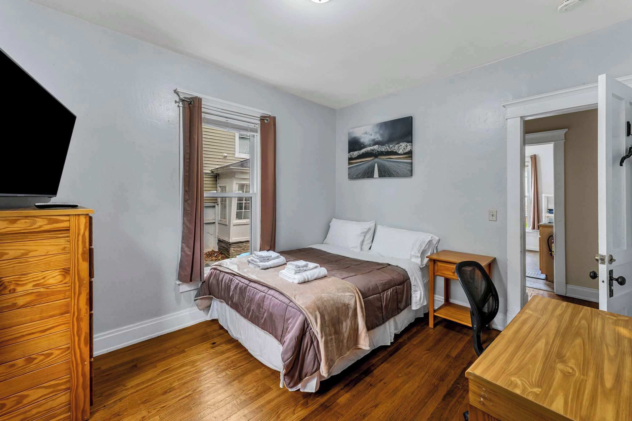 A bedroom with a bed, a desk, a chair, a dresser, a window with curtains, and a painting on the wall.