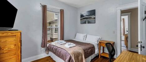 A bedroom with a bed, a desk, a chair, a dresser, a window with curtains, and a painting on the wall.