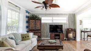 Un salon avec un canapé blanc, un meuble en bois, une télévision et un ventilateur de plafond.