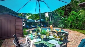 Une terrasse extérieure avec une table dressée pour le repas, entourée d’une végétation luxuriante et d’une piscine.