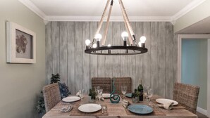 Une salle à manger avec une table en bois dressée pour quatre personnes, entourée de chaises en osier, d’un lustre et d’un tableau encadré accroché au mur.