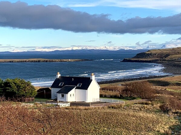 A white house with a black roof, situated near a coastline with a view of distant mountains and a cloudy sky.